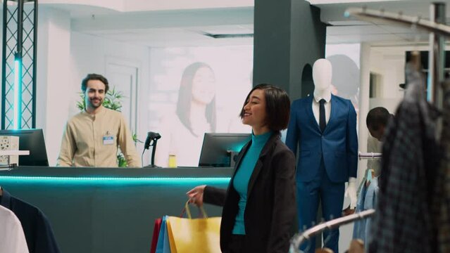 Female customer enjoying buying clothes from retail shop, doing dance moves in modern clothing store. Asian person feeling happy dancing in shopping center, holding shopping bags.