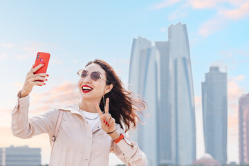 With a confident smile and a smartphone in hand, a girl captures a beautiful moment in front of the towering skyscrapers of Abu Dhabi.