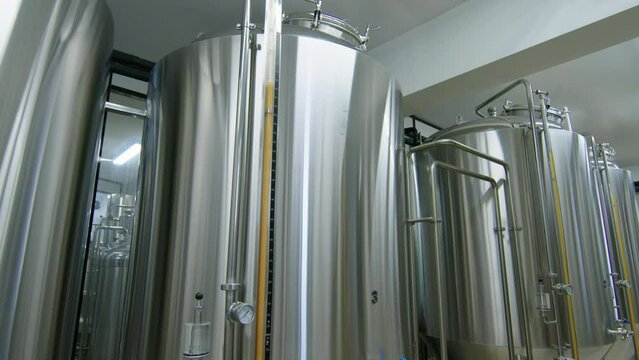 Medium travelling shot of row of large stainless steel bright beer tanks with pipes, sight tubes and gauges operating at small commercial craft brewery