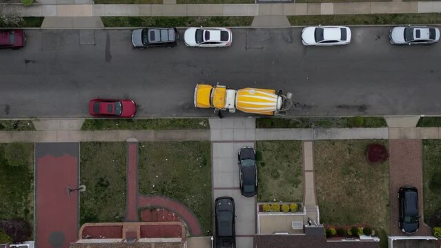 A Top Down View High Over A Yellow And White Cement Truck In A Residential Neighborhood On A Cloudy Day. The Drone Camera Is Tilted Straight Down And Boom Down Towards The Truck Mixing Cement.