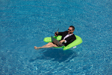 Funny business man in a business suit floating in the water in the pool. Remote summer work online. Crazy freelancer. Business and summer. Business man drink summer cocktail and using laptop in pool.