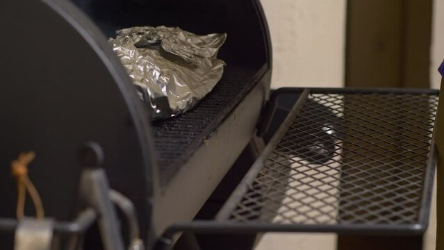 Man lifts BBQ smoker lid and uses tongs on foil wrapped ribs