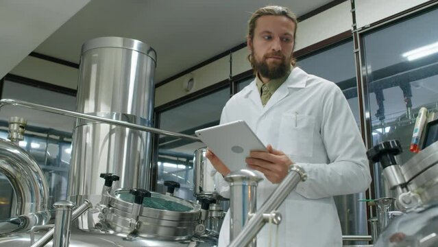Medium arc shot of young Caucasian male brewmaster in white coat standing on platform of brewhouse, observing beer brewing process through sight glass and filling checklist on tablet computer