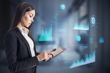 Attractive blonde businesswoman using tablet with glowing digital business interface with charts on blurry office interior background. Infographic and big data concept. Double exposure.