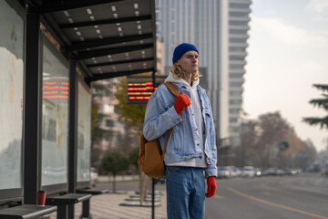 Upset backpacker student stylish guy citizen urbanite waiting bus on bus stop in downtown in spring...