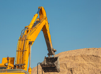 An orange excavator fills up the earth in the body of a construction dump truck. Excavator with bucket and dump truck, while working on the background of the blue sky close-up.