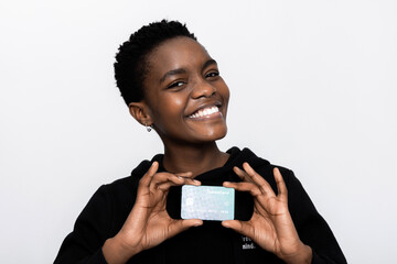 Delighted black african american woman in casual black hoodie smiling with white teeth at camera holding new modern credit card in hands standing over white background in studio isolated.Easy payment.