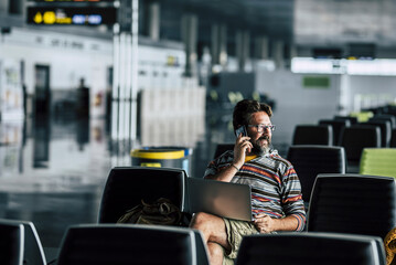 One travel man sitting at the airport gate waiting room and talking at the phone alone with laptop on legs. Concept of flight delay or cancel. Passenger tourist wait inside station. Traveler problem