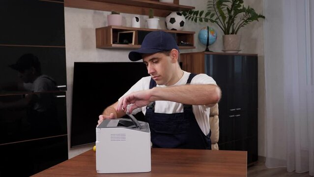 Close-up of a handyman in overalls, a white t-shirt and a cap with a screwdriver in his hands repairs the printer in the office or at the client's home. Call a home repairman.