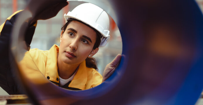 Latin engineer wears safety helmet work in heavy metal engineering factory. Young female hispanic worker in hardhat hard hat checking production machinery equipment in metalwork manufacturing facility
