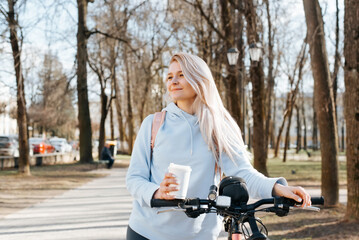 Pretty young woman with bicycle and cup of drink in park on walk, looking away outdoors. Relaxation, active rest