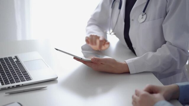 Doctor and patient sitting at the white desk near flair window in clinic. Unknown female physician wearing a white coat uses tablet computer for filling up medical record form. Medicine concept