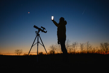 Astronomer looking at the starry skies with planets, falling stars and Moon eclipse with a telescope while using modern smartphone.