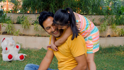 Urban girl child hugging her young dad from behind while he is reading a book. Medium shot of a handsome father enjoying with his daughter on a holiday outing in the park - happy family time
