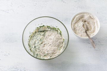 Glass bowl of mixed soft cottage cheese with herbs and green buckwheat flour on a light blue background, top view. Cooking homemade healthy cakes or bread
