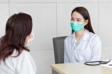 Asian female patient is attentively explaining her condition to a professional woman doctor in the clinic, both wearing masks to prevent infection at the hospital.