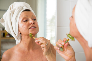 Mature beautiful woman doing facial massage with jade roller. Looking in the mirror in bathroom, Wrapped in a towel. Skincare routine and anti-aging treatment at home for older women.
