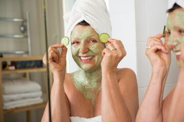 Mature beautiful woman applying green homemade DIY facial mask on skin. Looking in the mirror in bathroom, having fun. Wrapped in a towel. Skincare routine and anti-aging treatment for older women.