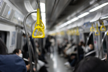 Selective focus of the yellow handle hanging on stainless steel handrail for standing passengers on an electric train with blurred passengers sitting on both sides of the electric train in background.