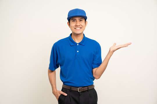 Happy delivery asian man in blue uniform standing on isolated white background. Smiling male delivery service worker pointing the finger to blank space. Courier and shipping service.