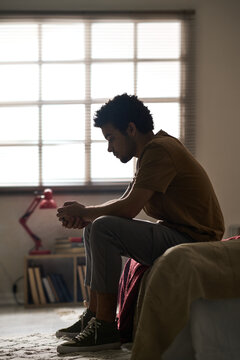Side view of young man staying alone with his thoughts sitting on bed in the room