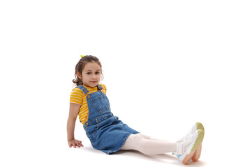 Horizontal full length portrait of Caucasian beautiful lovely 5-6 years old little kid girl in yellow t-shirt and denim sundress, sitting on isolated white background and looking confidently at camera