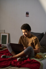 Vertical image of teenage boy using laptop while sitting on his bed and eating chips