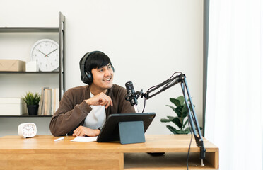 Young asian man host streaming podcast with condenser microphone work on tablet at small broadcast...