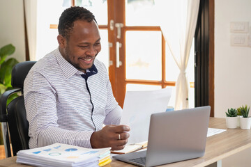 African American businessman of black descent sits smiling and doing financial reports and studying annual profit analysis An accountant checking the financial status of the company is in the office.