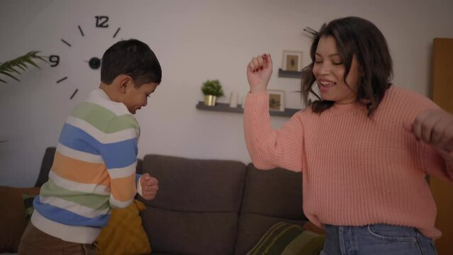 Latin mother and son dancing happily together in the living room. Excited and funny smiling little boy listening modern music with his young mom at home. Concept of childhood, family and sweet home
