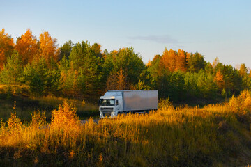 A truck of an unknown brand is driving on a road in an autumnal landscape bathed in the light of the setting sun. The autumn colors, the romance of a long-haul trucker, and the delivery goods on land.