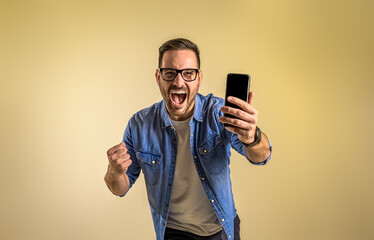 Portrait of cheerful male entrepreneur with smart phone screaming ecstatically and pumping fists. Young man reading message and celebrating success over beige background