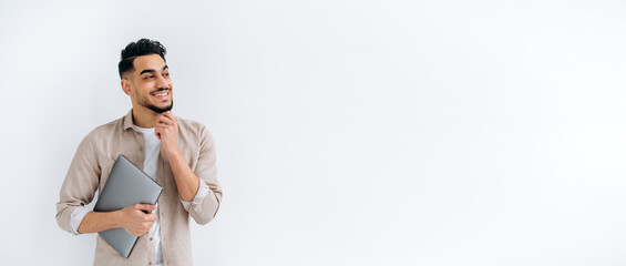 Panoramic photo of handsome arabian or indian guy in stylish casual wear, freelancer or student, holding a laptop, stand on white isolated background, looking away, smiling happily. Copy-space concept