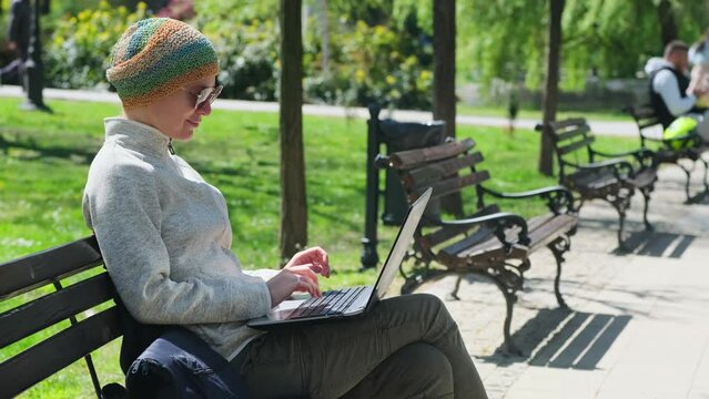 Caucasian woman in casual clothes and sunglasses typing on the laptop while sitting on the bench in a public park in sunny day. Female freelancer using her computer outdoor.