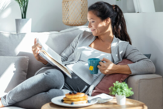 Beautiful relaxing pregnant woman reading a book while cup of tea lying on sofa at home