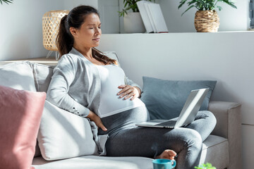 Worried pregnant woman with a stomachache working with laptop sitting on sofa in the living room at...