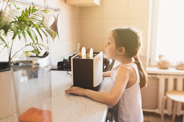 Funny caucasian girl 6 years old in a home kitchen looks with surprise at a toaster with bread on a working kitchen tabl, selective soft focus. - obrazy, fototapety, plakaty