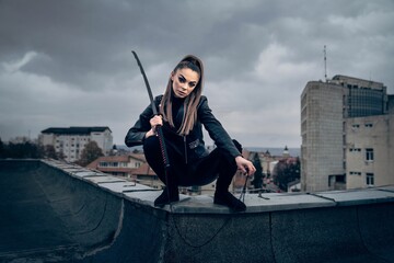 Cool female ninja warrior in leather outfit wielding a Japanese sword on the top of a building