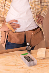 Crop carpenter standing with hands on waist near workbench with hammer nails and wooden blocks while working in workshop