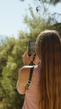 A young woman in a pine forest takes a phone view of the mountains. Sunny weather.Vertical video Social Media