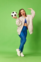 Portrait of beautiful young girl wearing sport wear holding ball and smiling over green studio background. Girls football