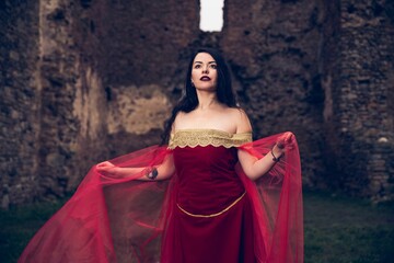Young beautiful female in medieval red dress posing near old ruins