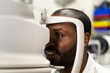 A dark-skinned man is being evaluated in a clinic using a tonometer to measure eye pressure. Eye...