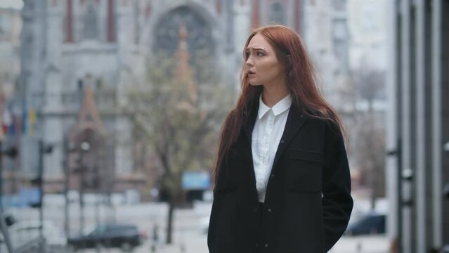Upset red-haired girl in black coat standing on the urban background. Outdoor video of lonely woman