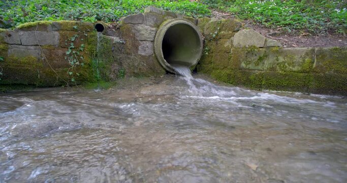 Small water stream running out of an industrial-size concrete pipe in a forest in Europe. Daytime, real time, no people, wide angle