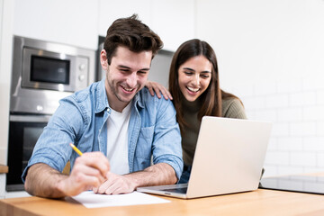 Fototapeta na wymiar young caucasian couple planning budget together at home writting notes to paper and looking a laptop. two cheerful young adults sitting on the kitchen. finance concept