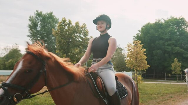 Girl in an equestrian outfit enjoying riding horseback along the trail, smiling and petting her beautiful red horse, handheld shot.