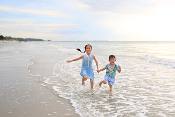 Happy Asian girl kid and little boy having fun running on tropical sand beach at sunrise. Happy family sister and brother enjoy in summer holiday.
