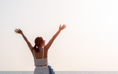 Back of Asian woman with sunglasses, happy raise hands up pose on the beach in summer vibes. Happy female in white tank top looking up to sky on sea and open sky with copy space, Holiday vacation.