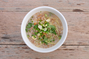 Rice porridge with minced pork, garlic, coriander, chili, spring onion and ginger in white bowl against wood table background. Top view.